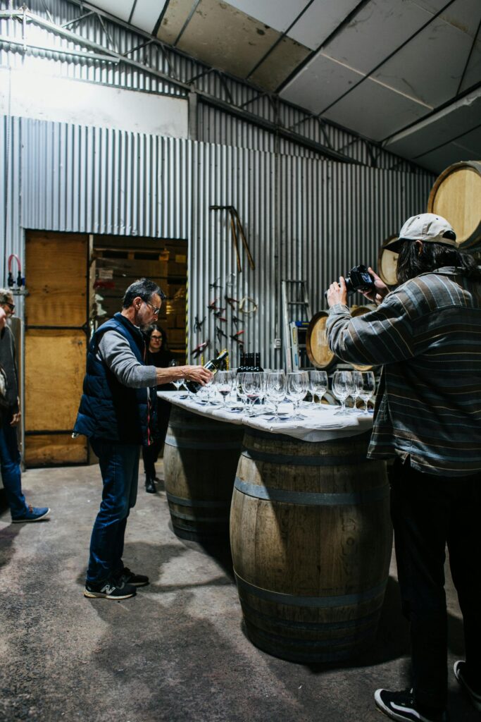 Group enjoying wine tasting at a rustic winery with barrels and glassware.