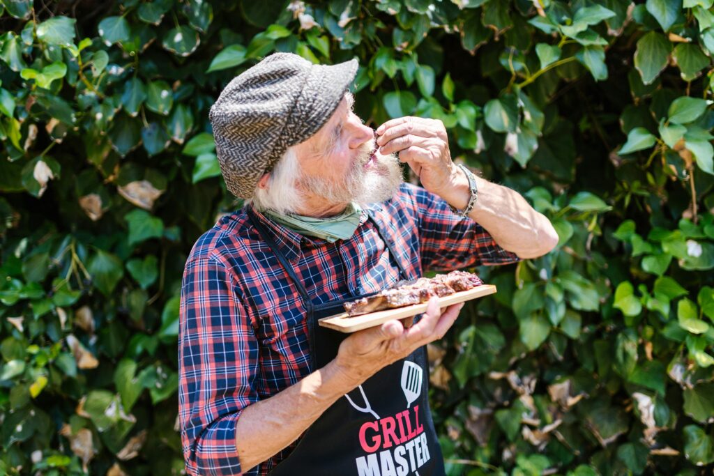 Elderly man in apron savoring grilled meat outdoors against a leafy backdrop.